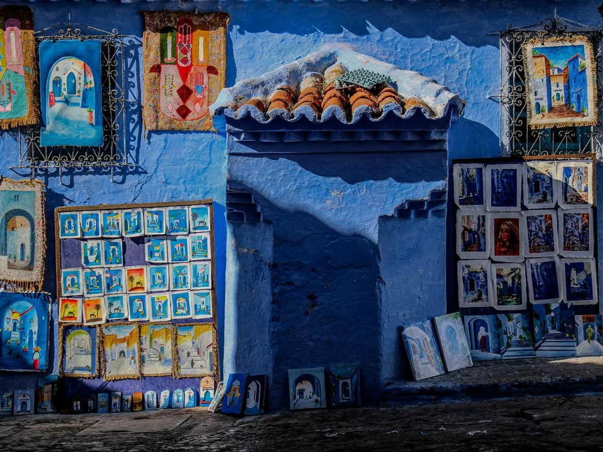 Local paintings and woven crafts hung for sale against a blue medina wall in Chefchaouen